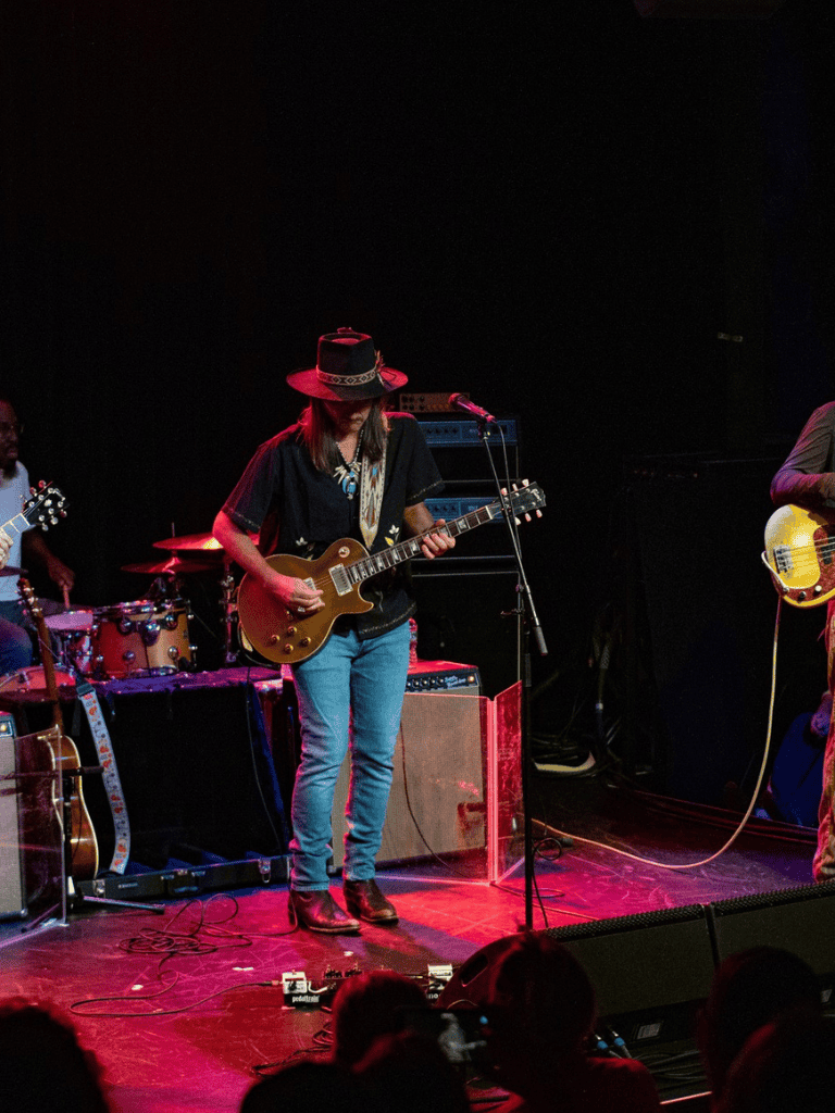 A female guitarist performing live on stage with band members, musical equipment, and vibrant stage lighting.
