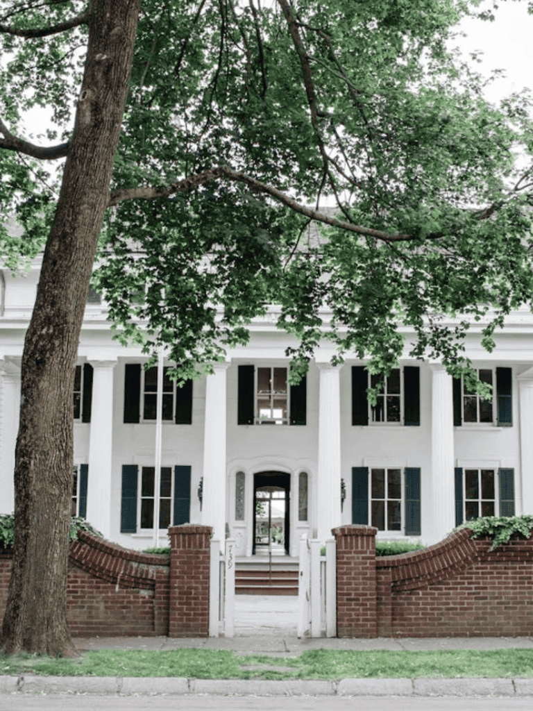 Grand White House with tall columns and black shutters in a lush green setting.