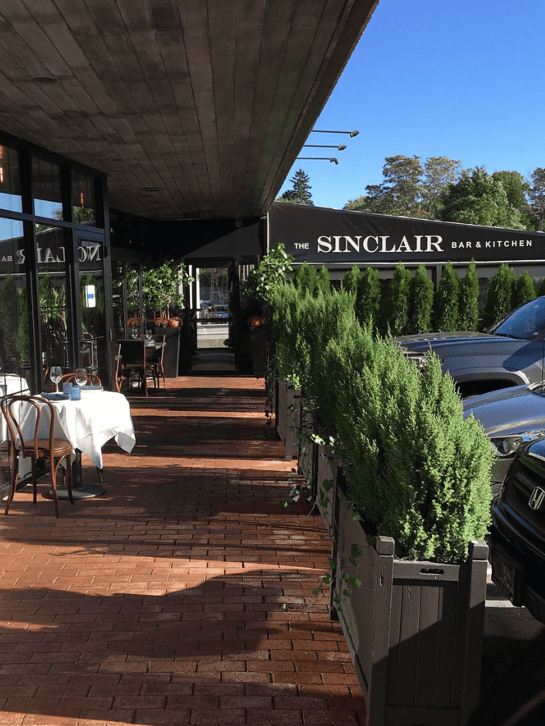 A beautiful outdoor dining area at Sinclair Bar & Kitchen with greenery and parked cars under a sunny sky.
