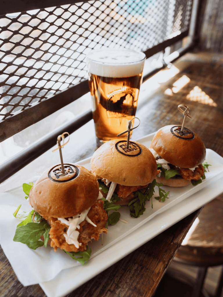 Fresh chicken sandwiches with greens served on a long white platter, accompanied by a cold beer at a casual eatery.