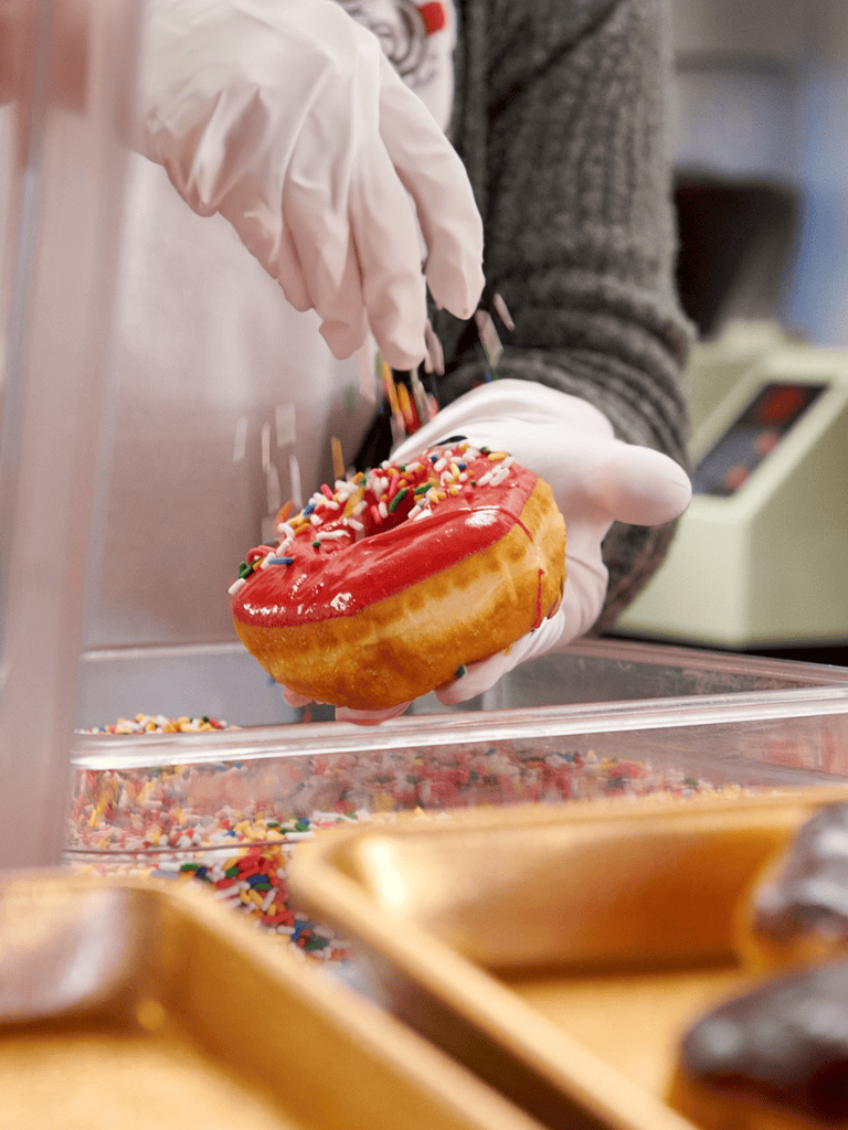 Donut with red icing and sprinkles being decorated in a bakery.
