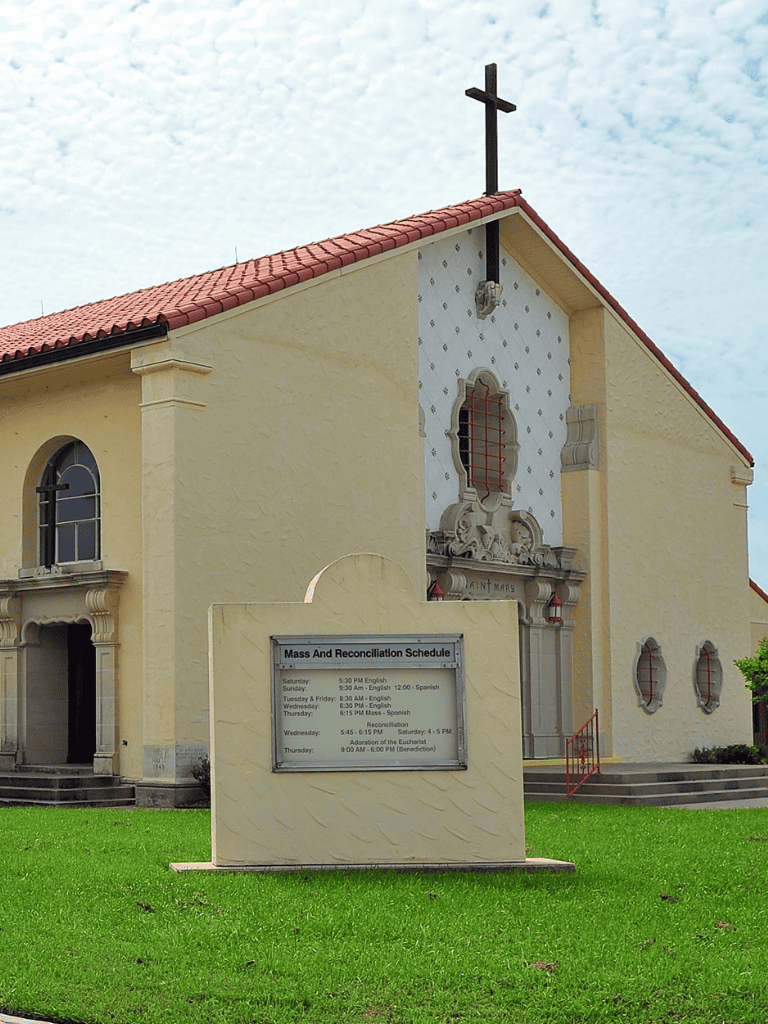 Quiet church with Mass schedule and a cross on the roof in a peaceful setting.