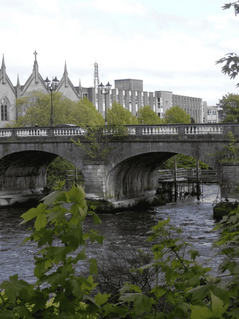 Quiet river flowing under historic stone bridge in downtown city surrounded by trees.