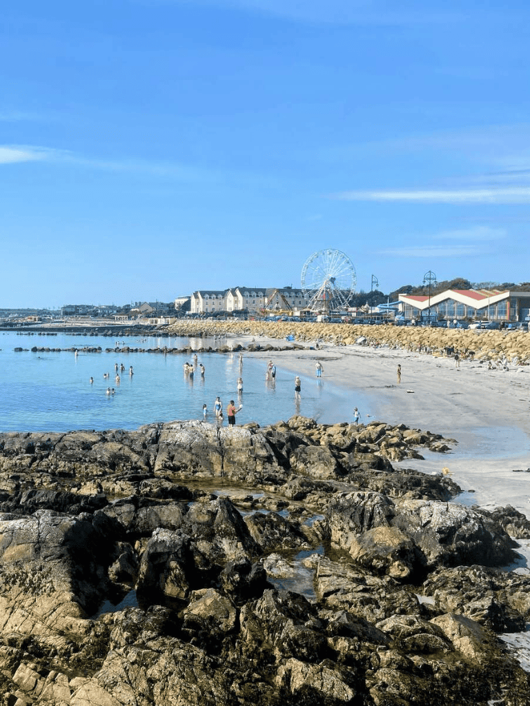 Relaxing beach scene at the seaside with rocks, people, and a Ferris wheel in the background, perfect for travel and tourism.