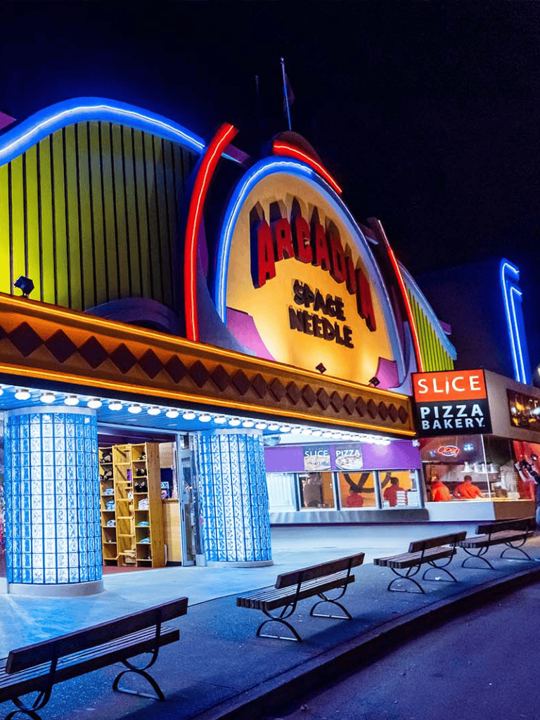 Bright neon-lit arcade with pizza bakery and seating area at night.
