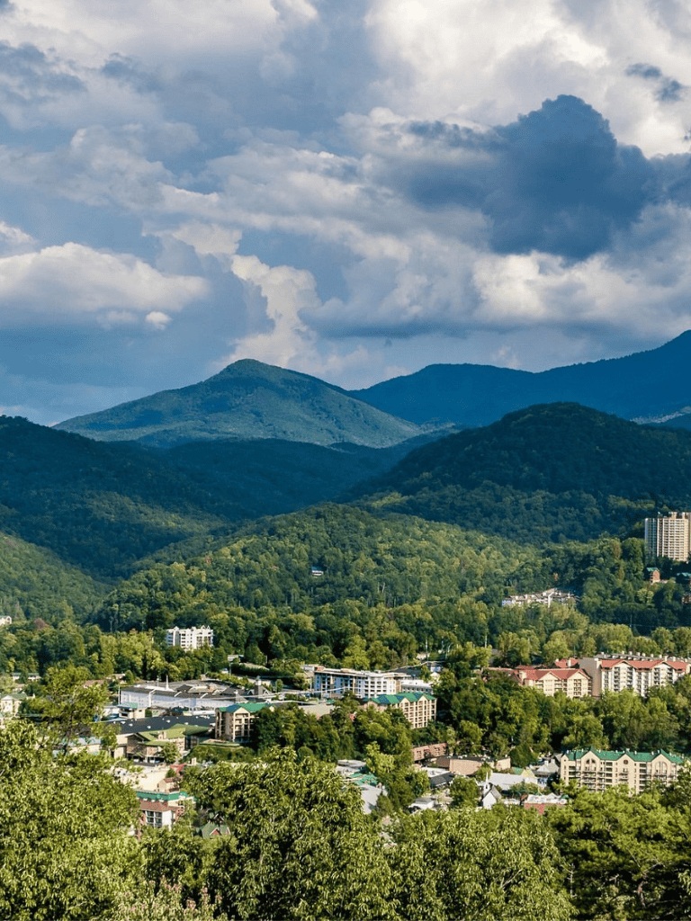 Vast mountain landscape with lush green trees and residential buildings under dramatic cloudy sky.