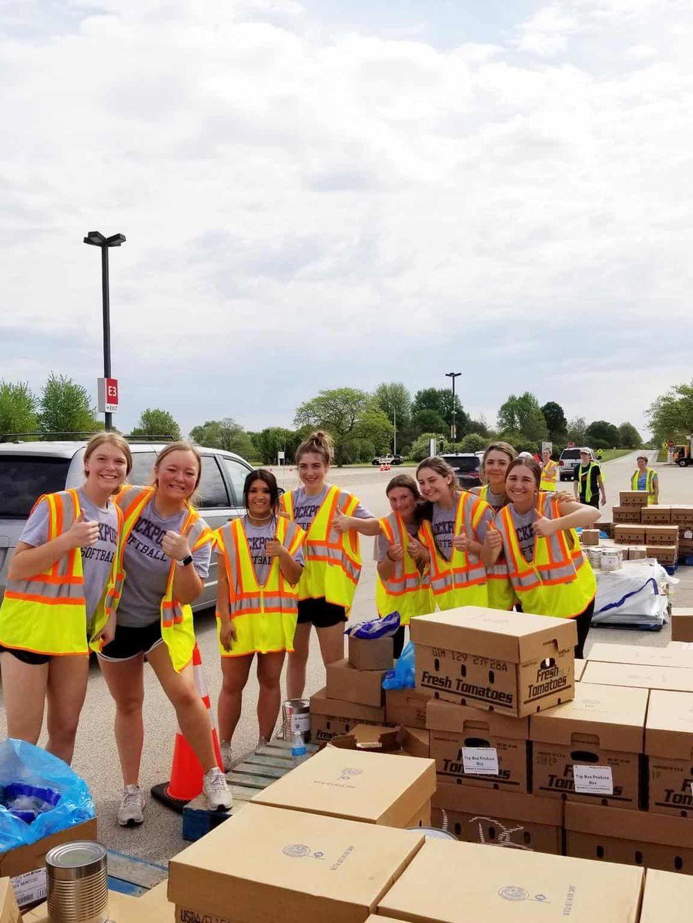 Volunteers at food distribution event wearing safety vests, smiling, and giving thumbs up.