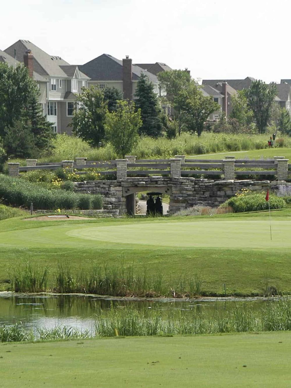 Serene golf course with a stone bridge and green fairways, surrounded by residential houses and lush trees.