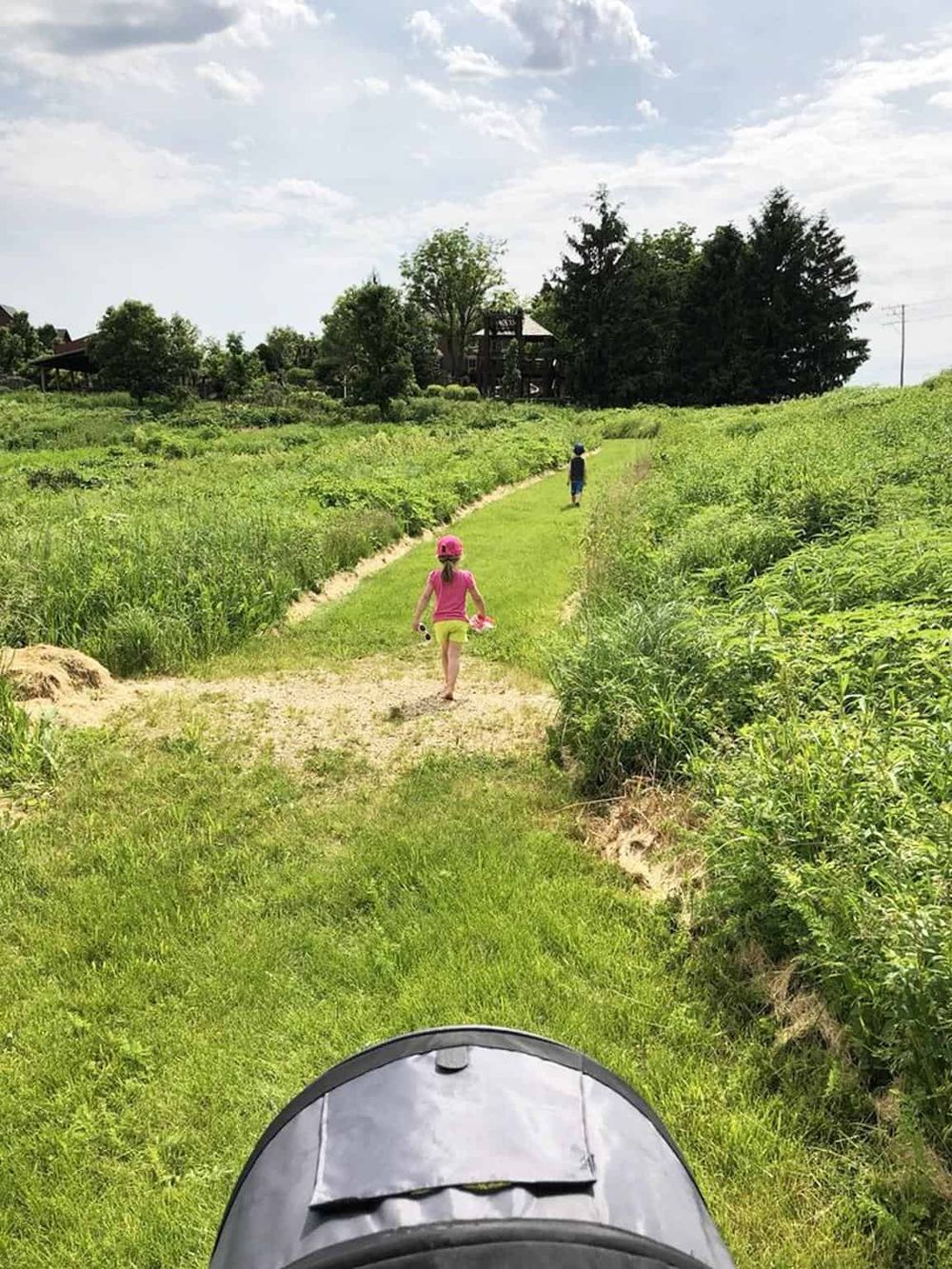 Child walking on a grassy trail in nature with a stroller in the foreground.