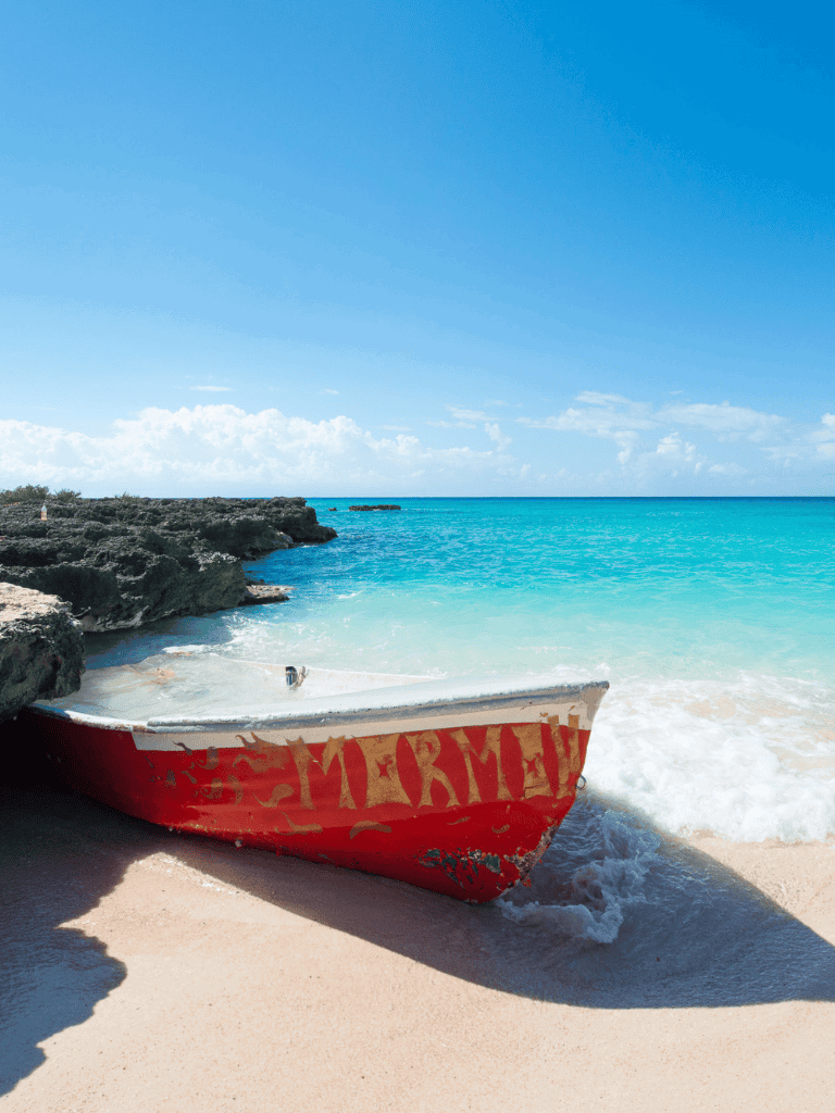 Quiet beach with turquoise water, rocks, and an old boat on sandy shore under clear blue sky.