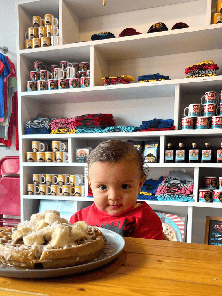 Banana waffles with ice cream served with kid at dessert shop, cozy interior featuring colorful merchandise and branded mugs.