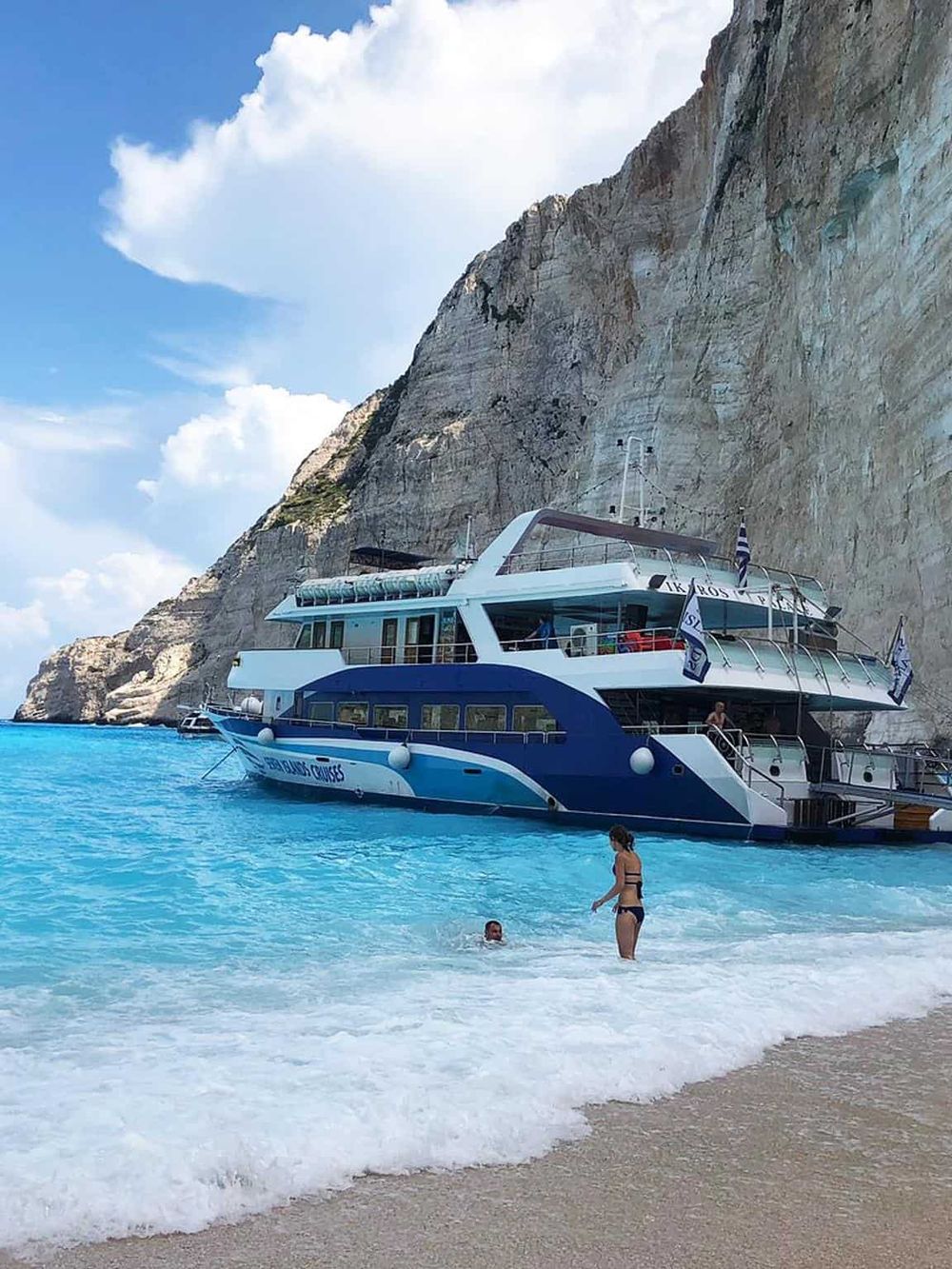 Luxury yacht docked at beach with people swimming and scenic cliffs in background, perfect for boat cruises.