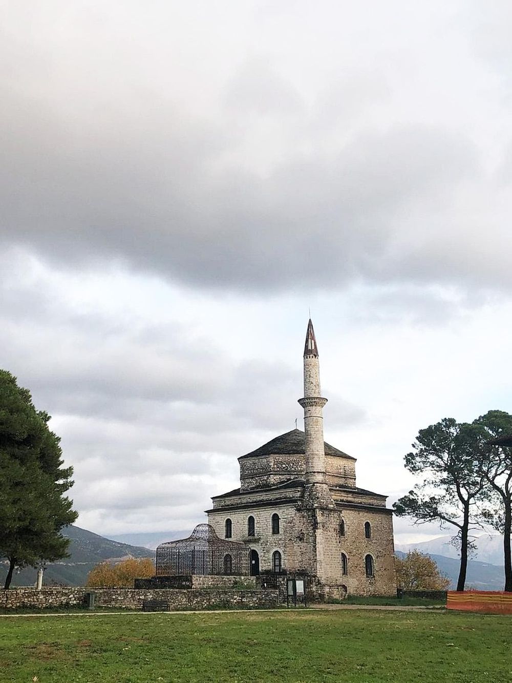 Ancient stone mosque with minaret, historic landmark, surrounded by greenery and cloudy sky.