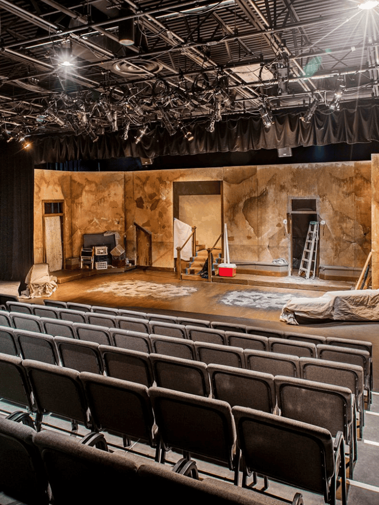 Stage theater set with wooden and painted backdrops, lighting, and seats ready for performance.