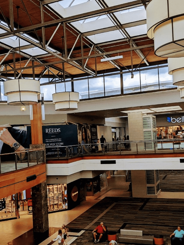 Bright indoor shopping mall with multiple floors, modern lighting, and visible stores and shoppers.