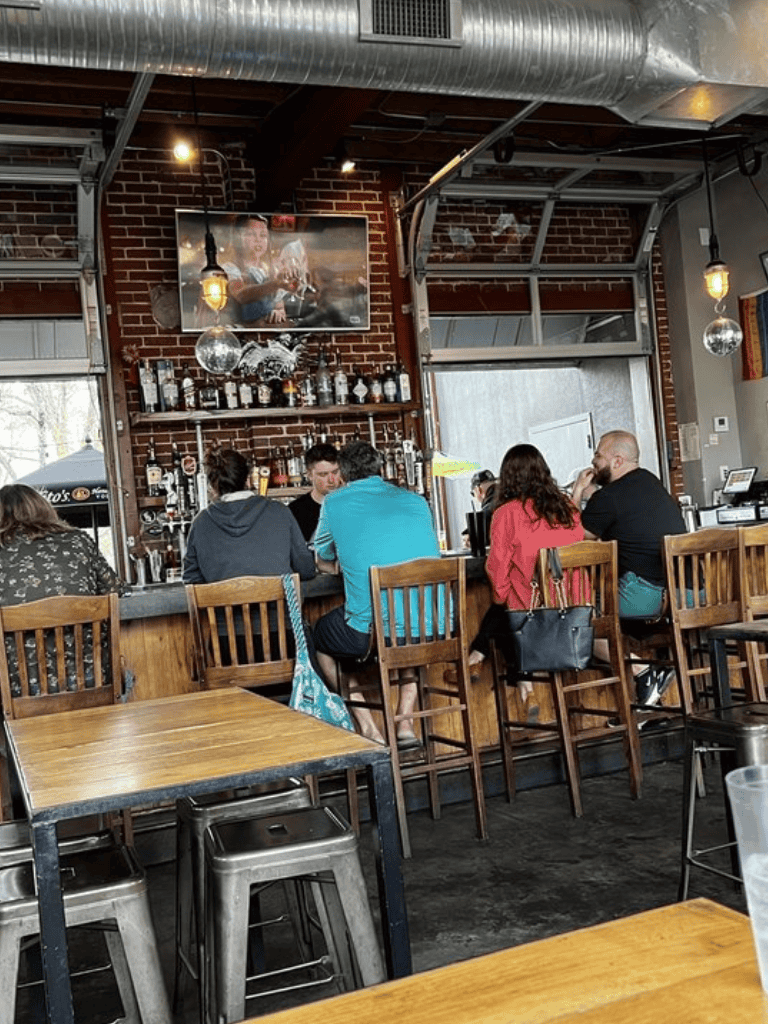 Bright bar interior with patrons socializing and watching sports on large TV screen.