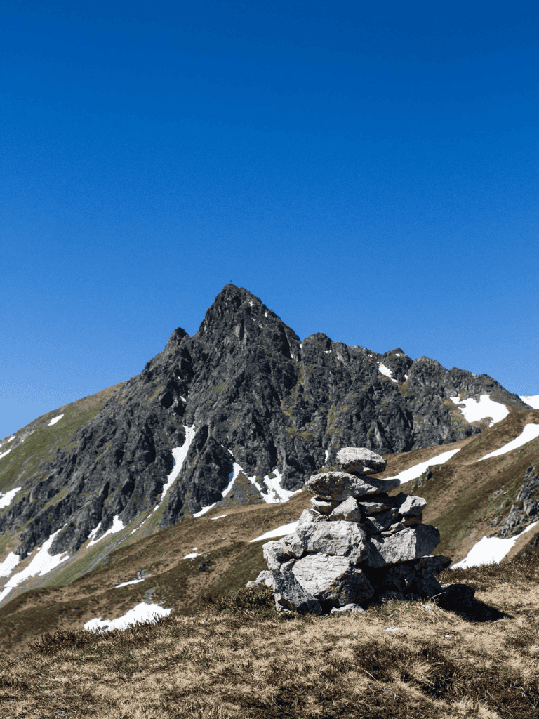 Stacked rocks cairn in mountain landscape with snow patches, clear blue sky, and rugged peaks.