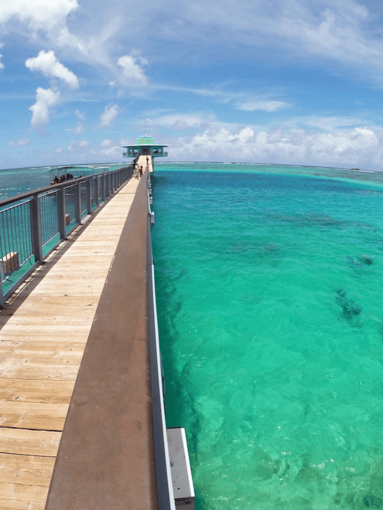 Bright turquoise ocean view with a wooden pier leading to a small building, under a partly cloudy sky.