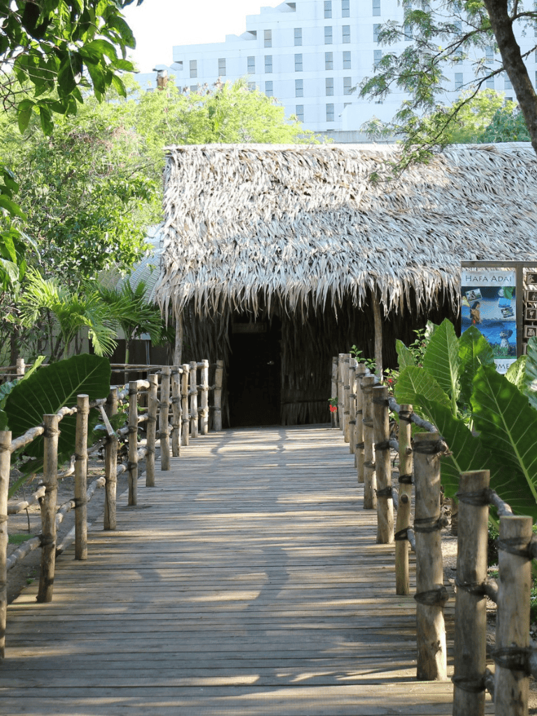 Thatched roof hut with wooden bridge, tropical greenery, and modern buildings in background.