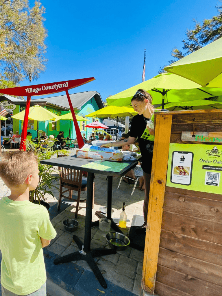 Open-air village courtyard with food vendor, children, and colorful umbrellas for outdoor dining and family fun.