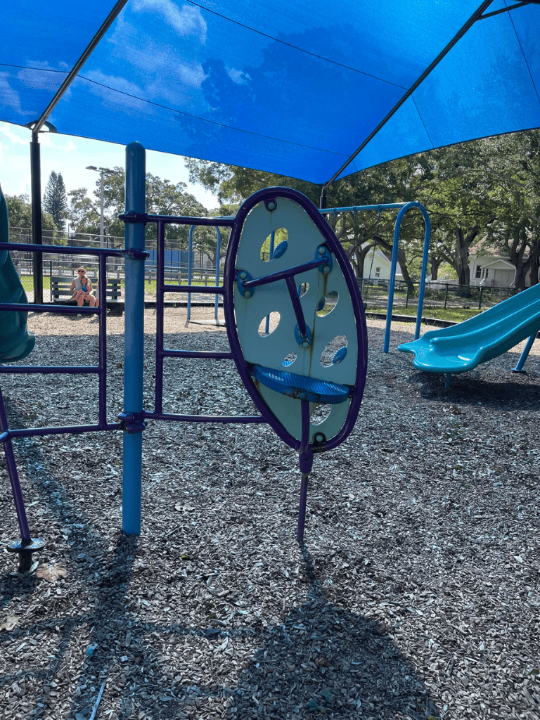 Bright blue playground structure under shade canopy with a slide, in a park setting with trees and a bench.