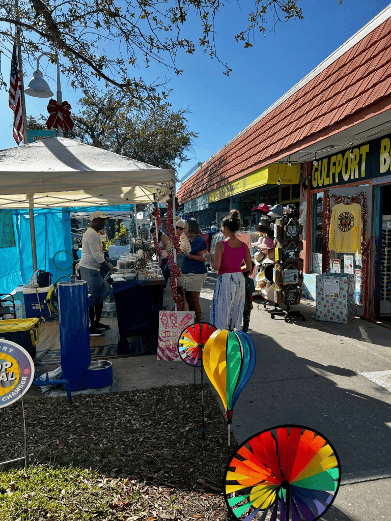Colorful outdoor market scene with stalls, shoppers, and decorative pinwheels on a sunny day.