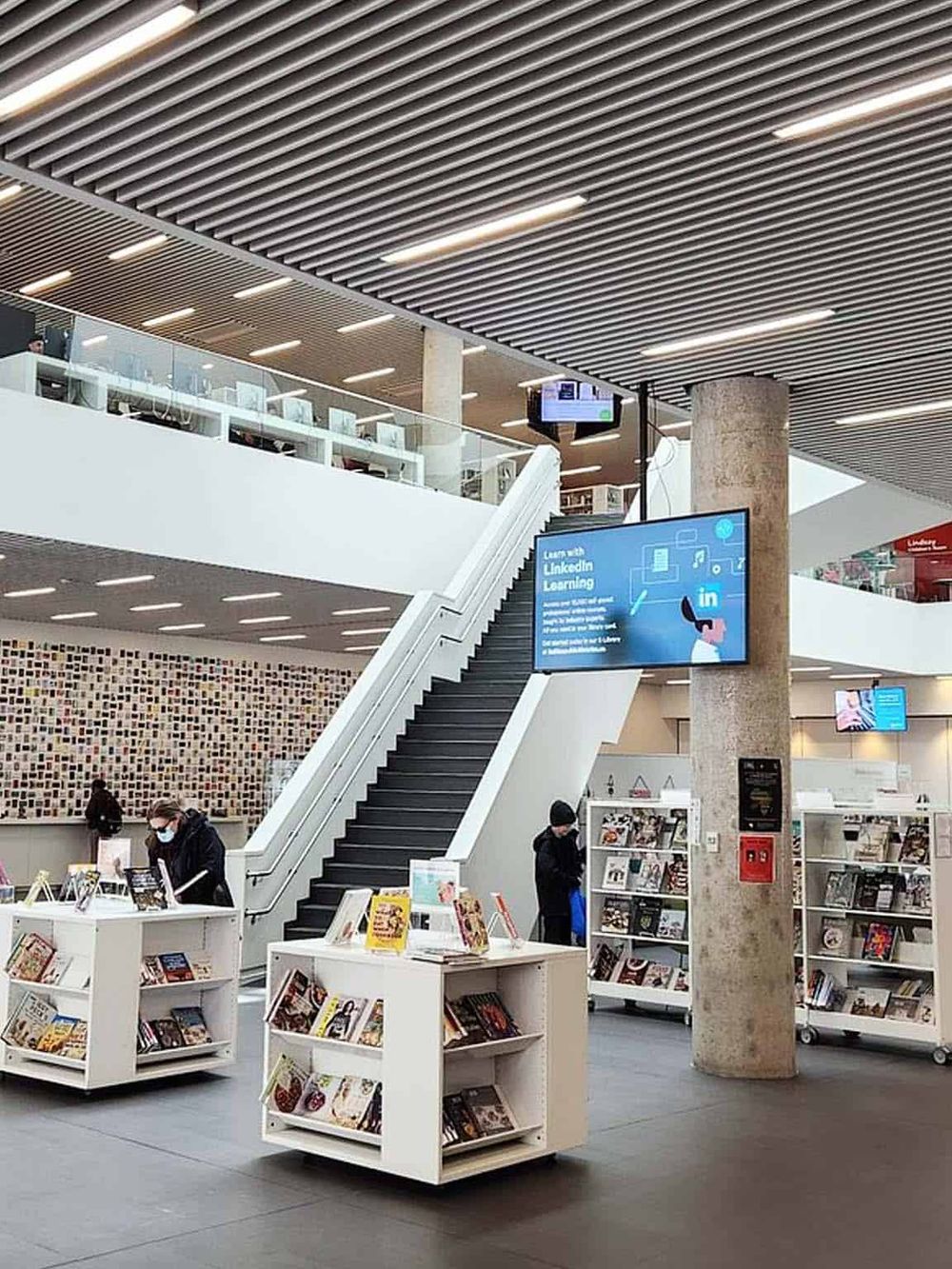 Modern library with bookshelves, digital screens, and an escalator in a contemporary interior setting.