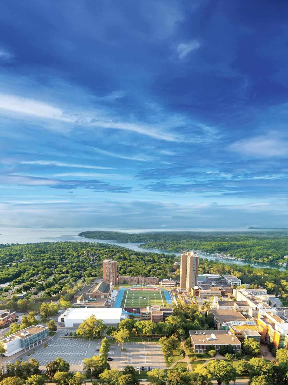 Aerial view of Quest for Directions campus with football field, tall buildings, lush greenery, and scenic river under a vibrant blue sky.