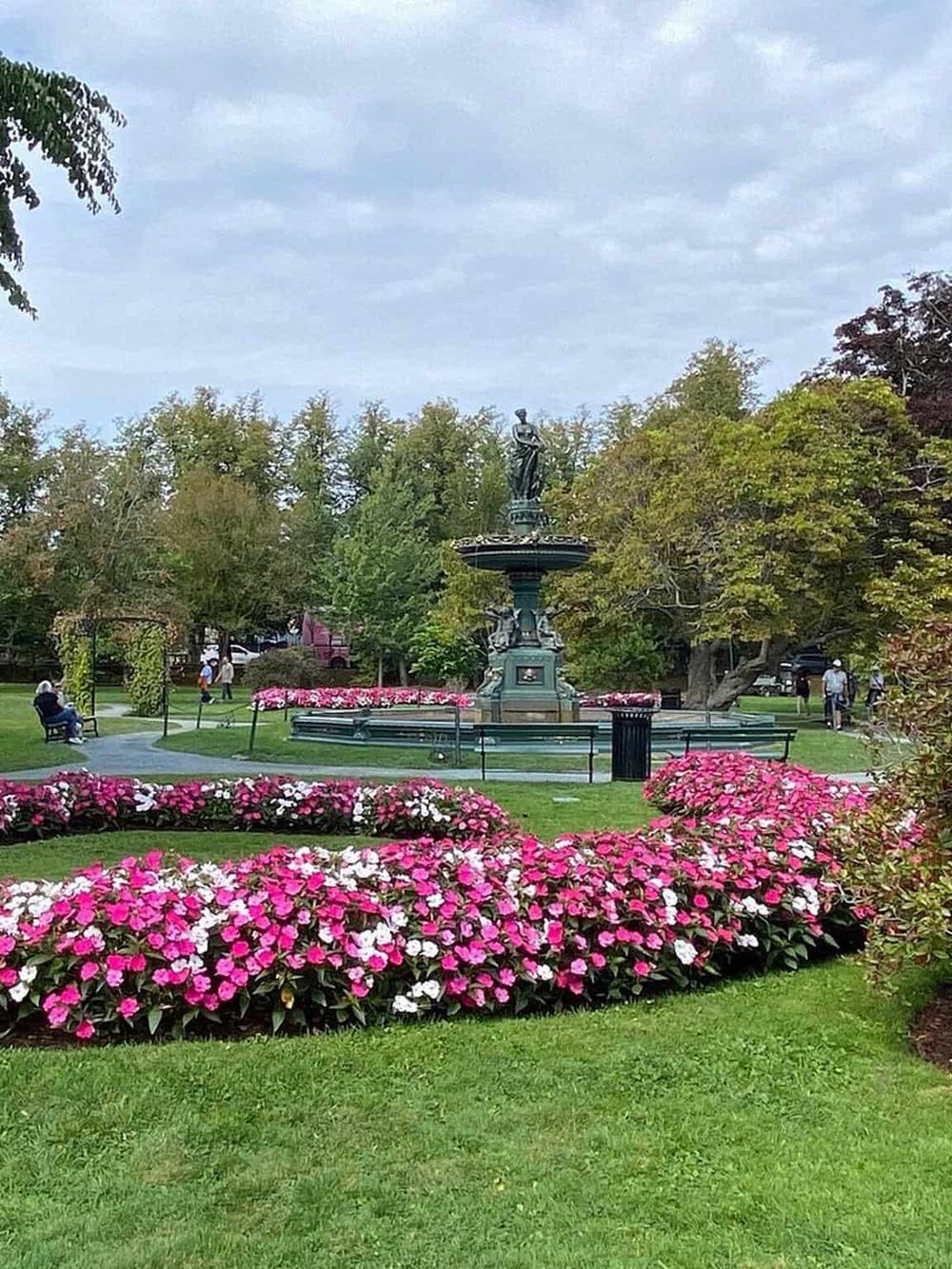 Beautiful park with a central fountain surrounded by vibrant pink and white flowers, lush green trees, and peaceful visitors.