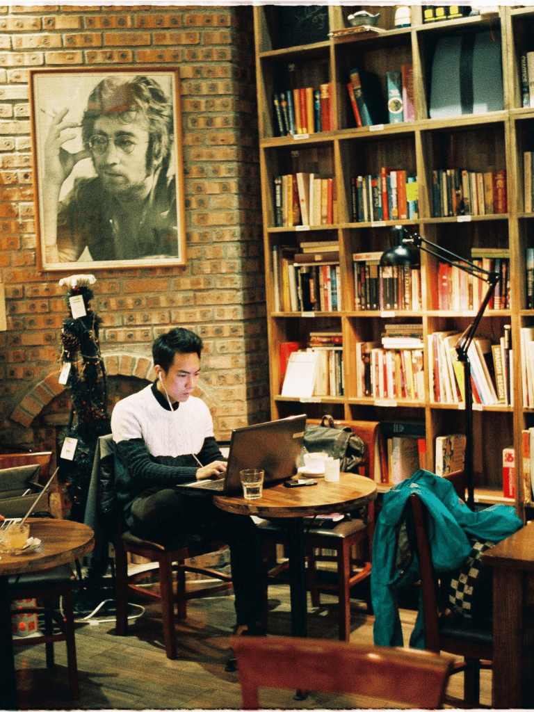 Cozy coffee shop with a young man working on a laptop, surrounded by bookshelves and warm brick decor.