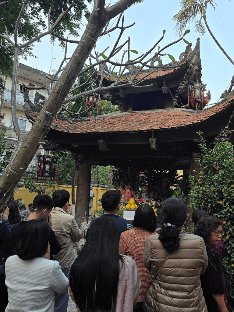 Devotees gather around a traditional Vietnamese shrine with a curved roof, offerings, and hanging red lanterns.
