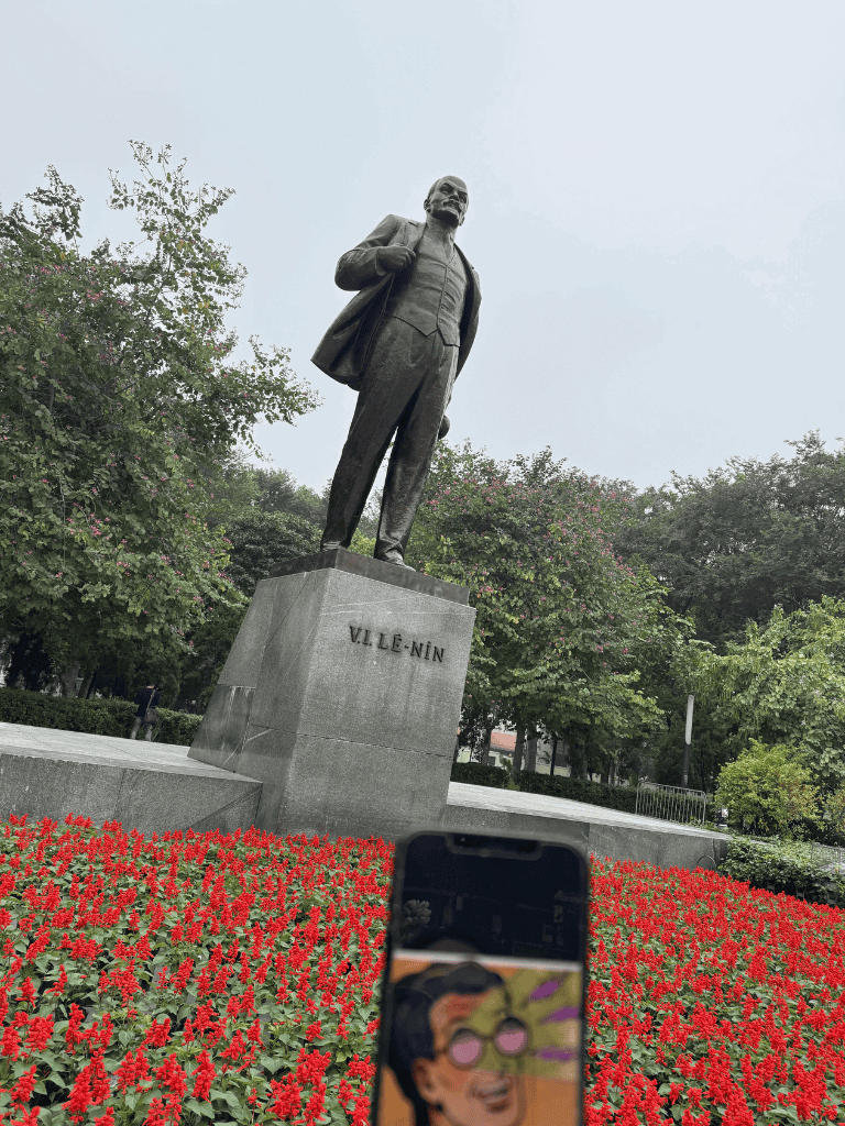 Statue of V.I. Lenin in a park with red flowers, greenery, and overcast sky.