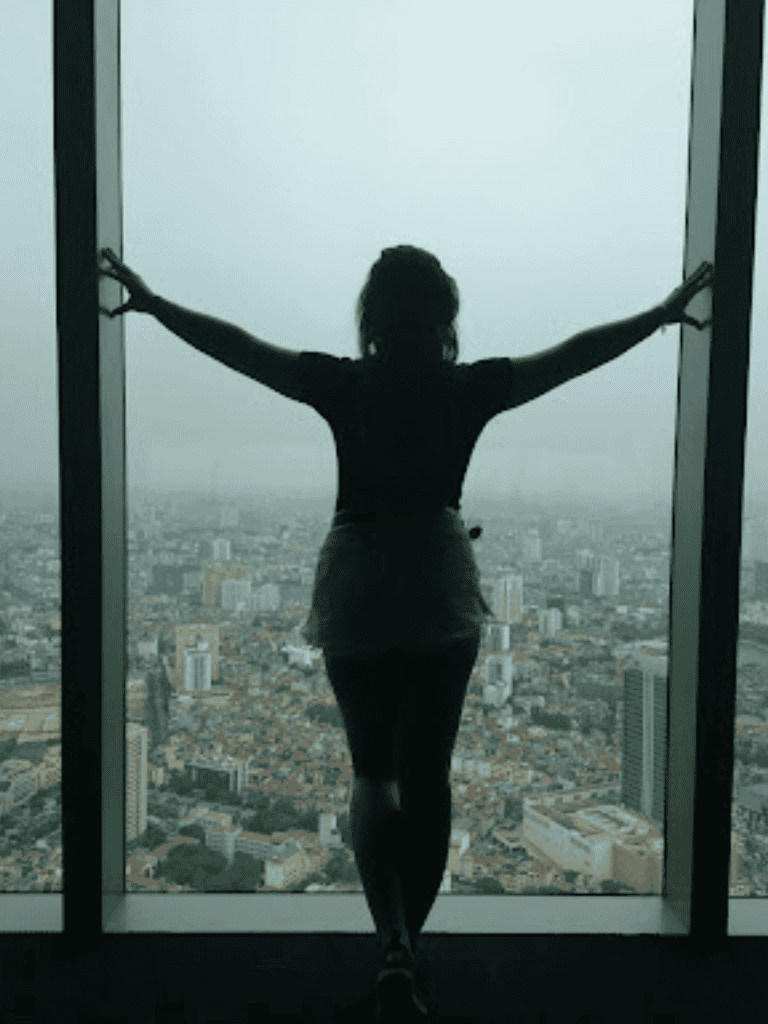 Vast city view from skyscraper window with woman standing in front, arms outstretched.