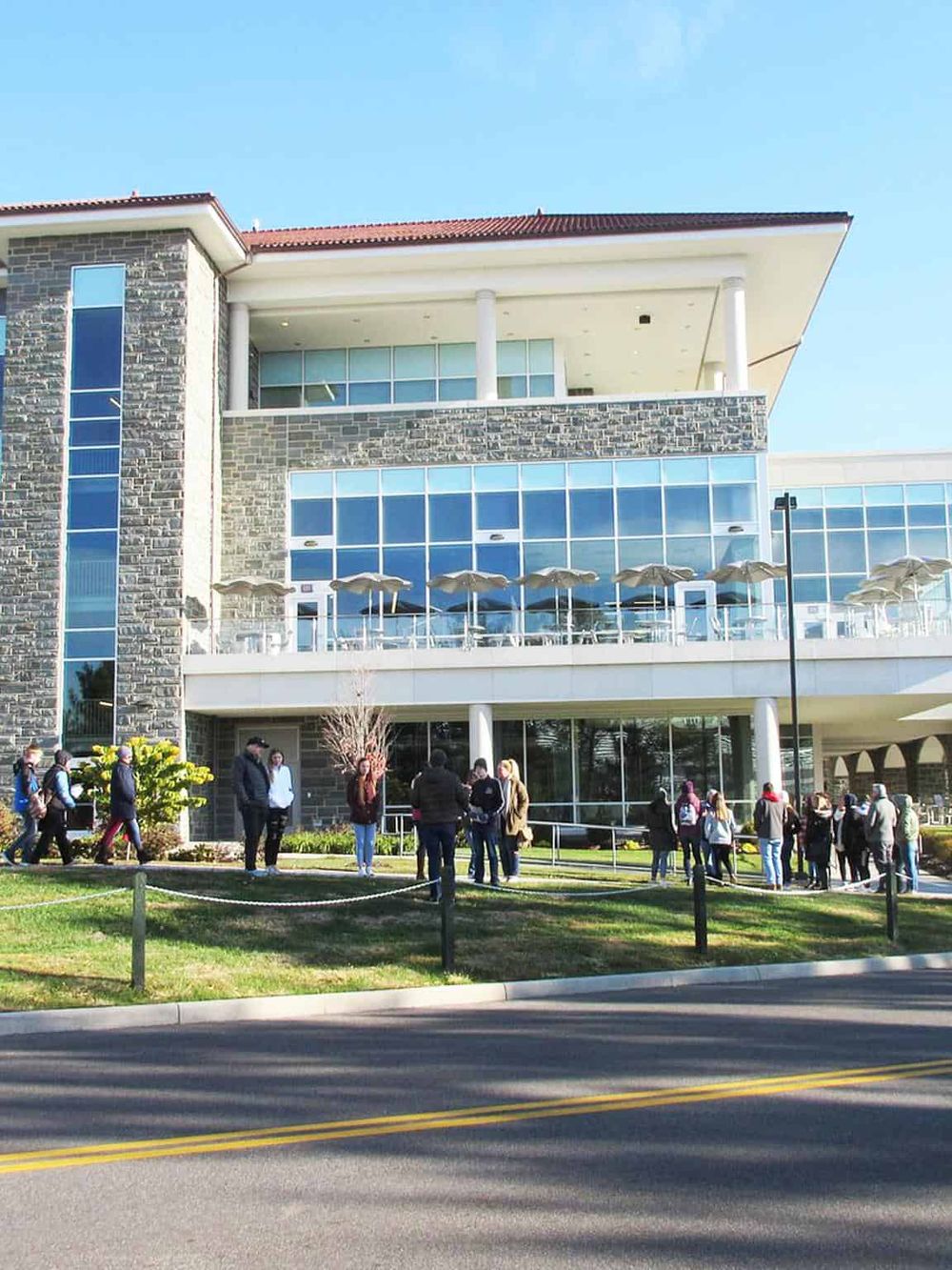 Modern multi-story building with glass windows, people waiting outside, and a clear blue sky.