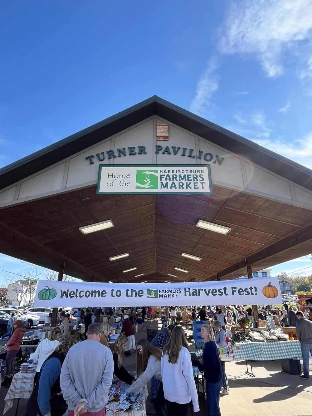 Colorful farmers market at Turner Pavilion with Harvest Fest banner under clear blue sky, lively outdoor community event.
