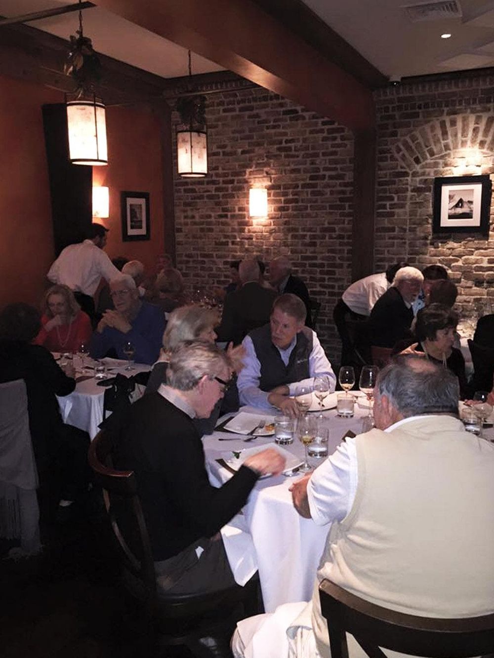 Cozy restaurant dining scene with elegantly dressed guests enjoying a meal in a warmly lit brick-walled venue.