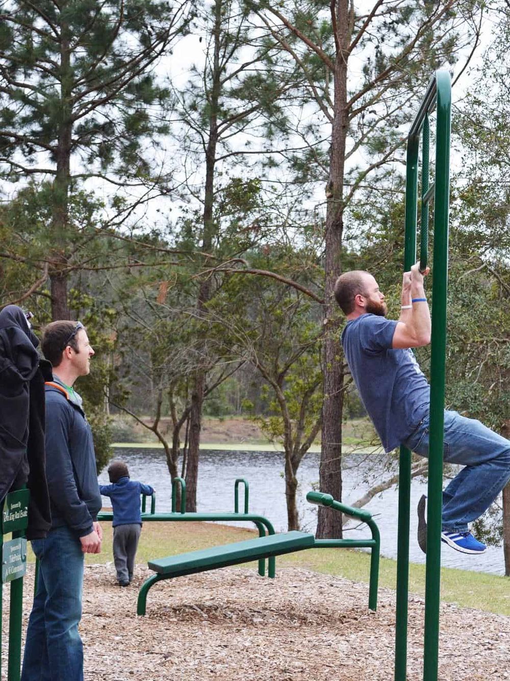Outdoors fitness at park with people using pull-up bars near lake and trees, promoting outdoor exercise and wellness.