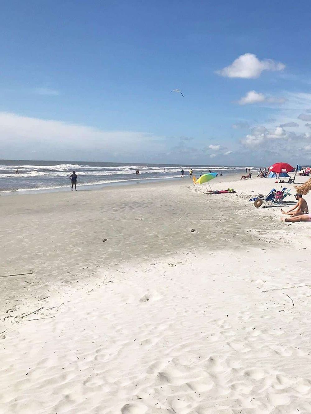 Soft sandy beach with umbrellas, beachgoers, and ocean waves under blue sky with scattered clouds.