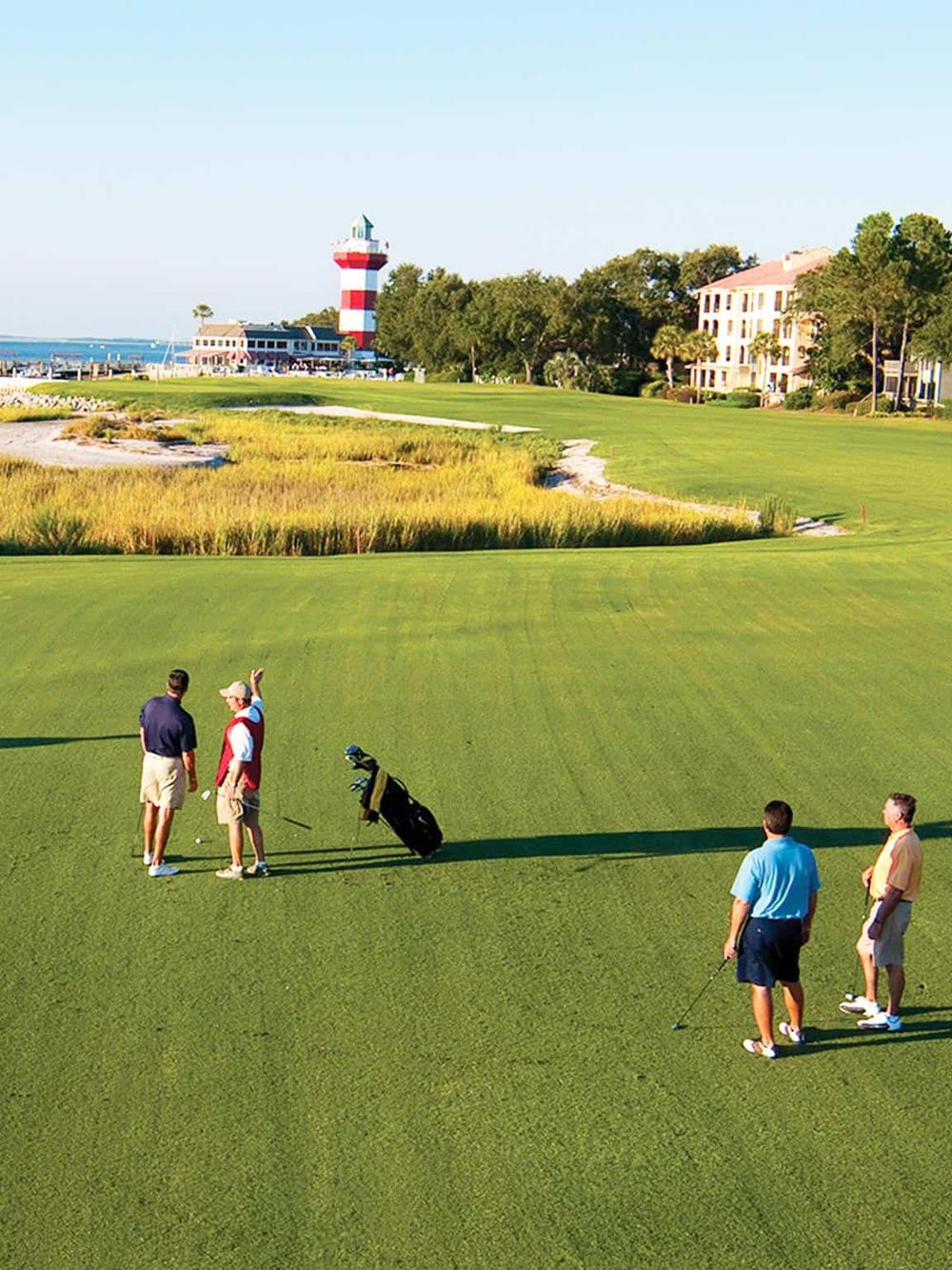 Bright golf course near waterfront with lighthouse, buildings, and people practicing putting, promoting golf and waterfront recreation.