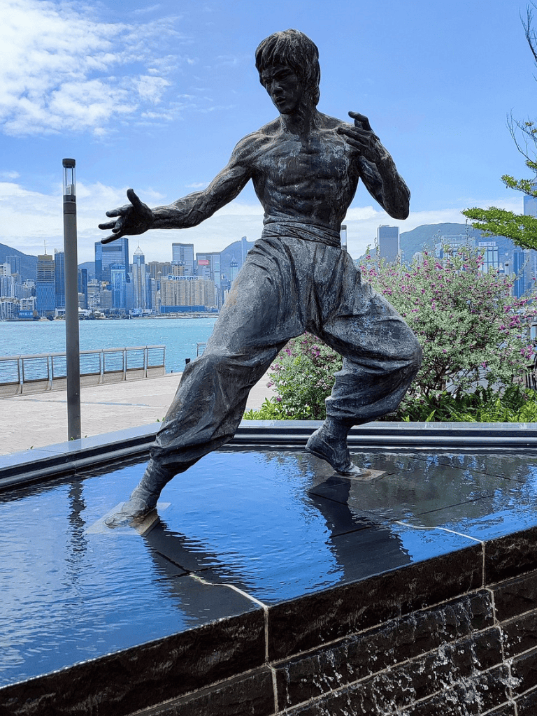 Dynamic Bruce Lee sculpture in Hong Kong waterfront with city skyline backdrop, symbolizing martial arts excellence.