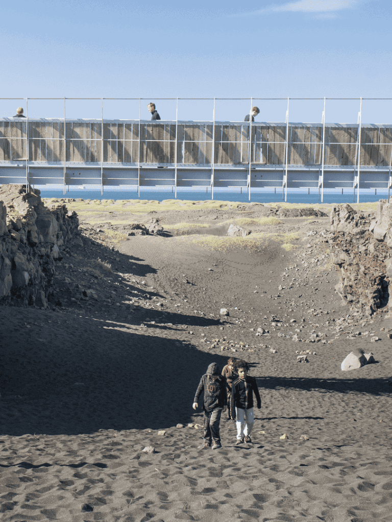 Amusement park or boardwalk with people walking on a bridge over rocky terrain and beach.