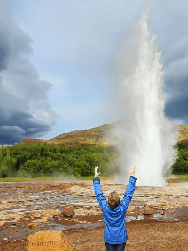 Vibrant person with arms raised in front of a stunning geyser eruption in Iceland, showcasing natural geothermal wonders.