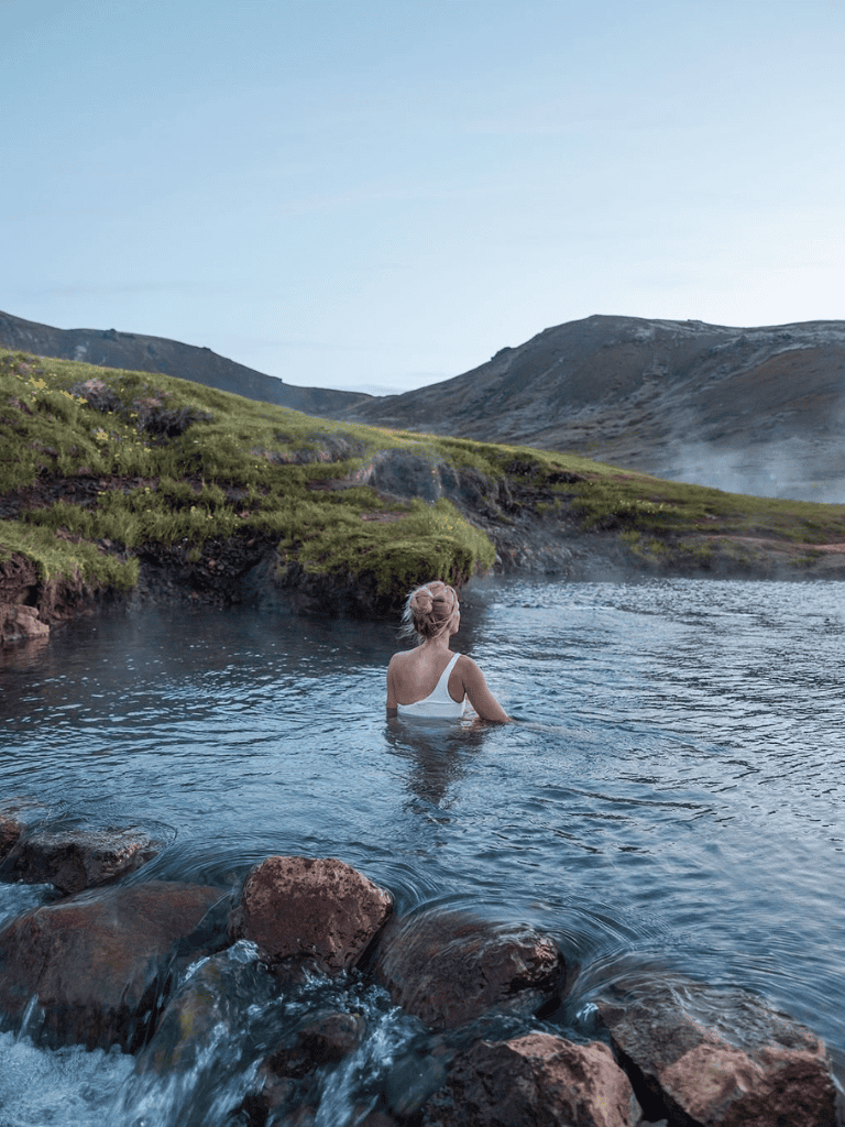 - Tranquil woman relaxing in geothermal hot spring surrounded by lush green mountains and rocky terrain.