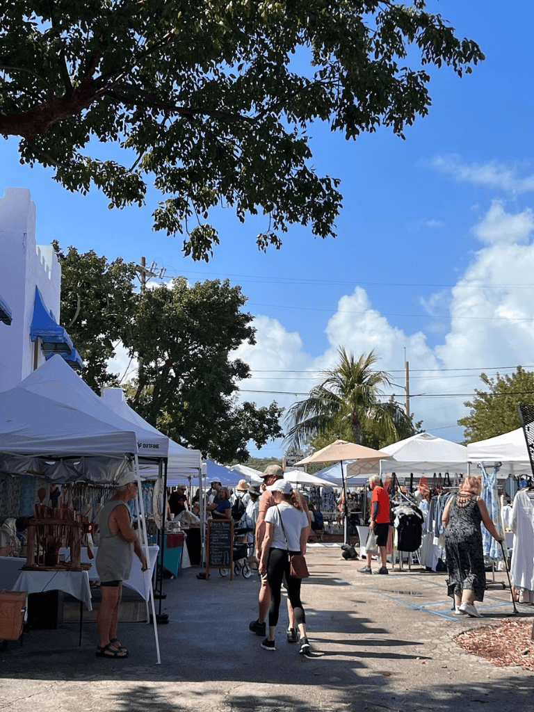 Vibrant outdoor market with white tents and shoppers, under blue skies in a tropical setting.