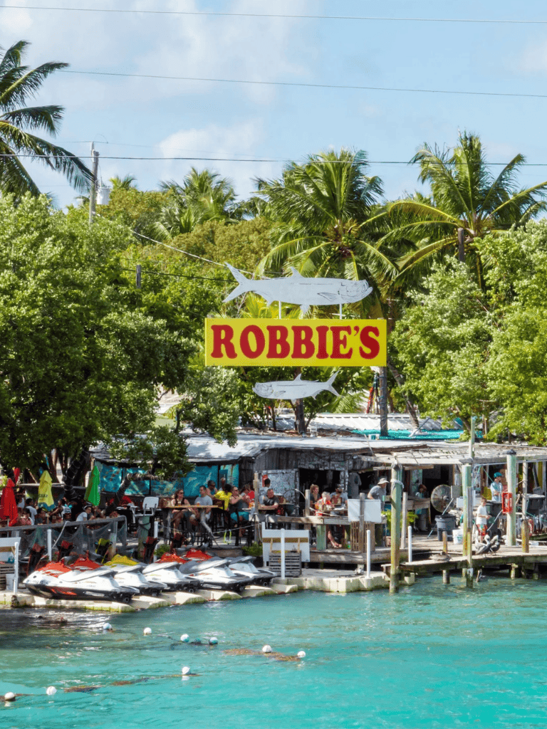 Bright tropical waterfront restaurant with outdoor seating, jet skis, and lush palm trees at Robbie's on Islamorada.