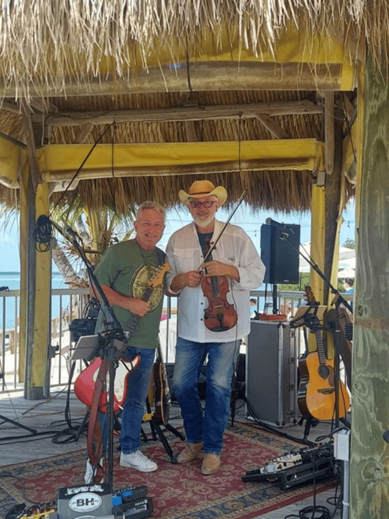 Guitarist and violinist performing at a tropical outdoor venue under a thatched roof.