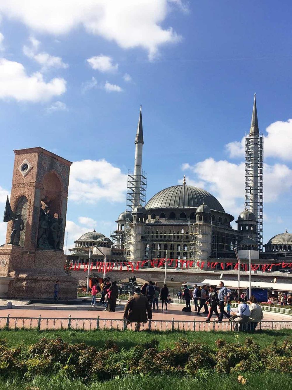 Historic mosque under construction with scaffolding, minarets, and a bustling square in front.