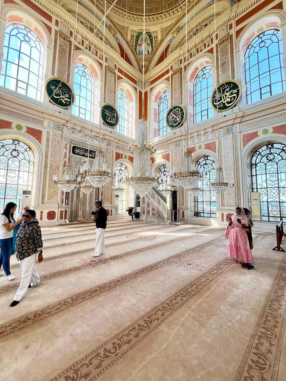 Intricate mosque interior with ornate chandeliers, large arched windows, and Arabic calligraphy decor.
