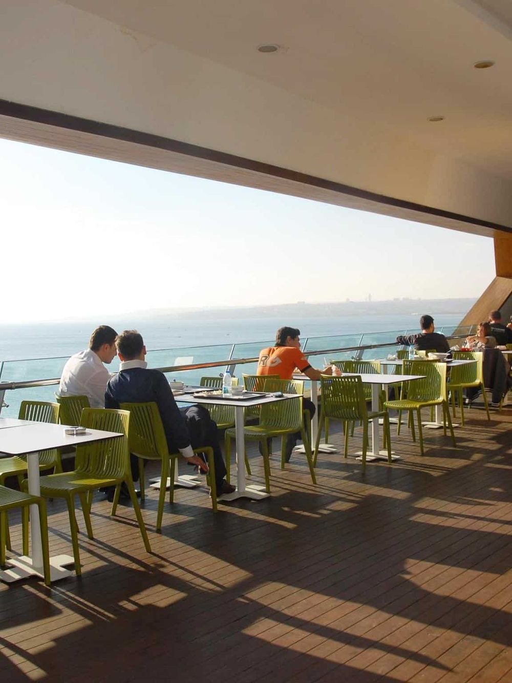 Panoramic ocean view from a modern, open-air restaurant balcony with seated diners relaxing in sunlight.