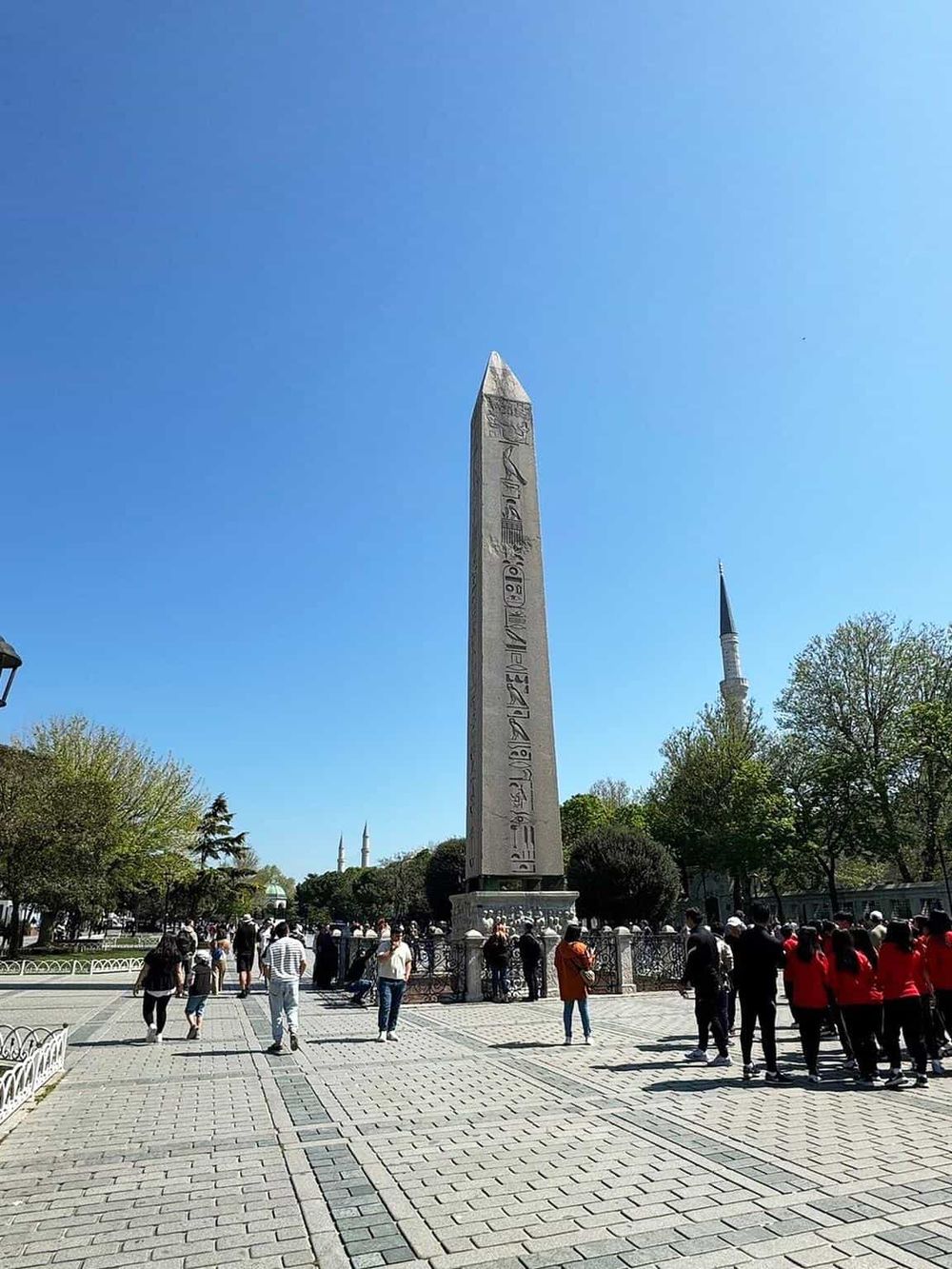 Ancient Egyptian obelisk in a lively park with visitors and historic structures behind, under clear blue sky.